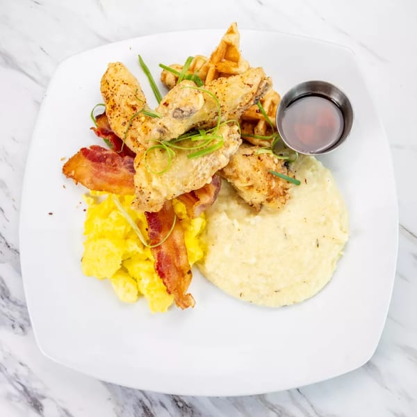 Fried chicken wings, waffle pieces, scrambled eggs, bacon, grits, and syrup on a white plate.