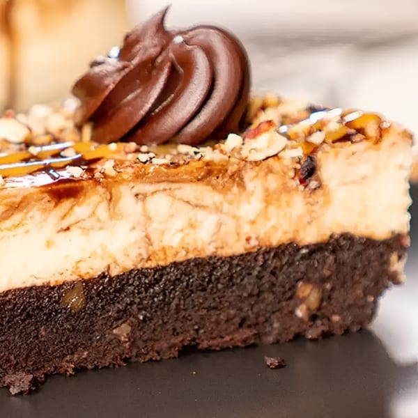 Close-up of a cheesecake slice with a brownie base, caramel, nuts, and chocolate swirl.
