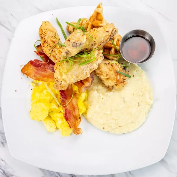 Fried chicken wings, waffles, scrambled eggs, bacon, grits, and syrup on a white plate.