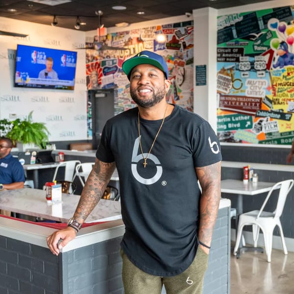 Man in a black shirt and cap smiling in a restaurant with a colorful collage wall.
