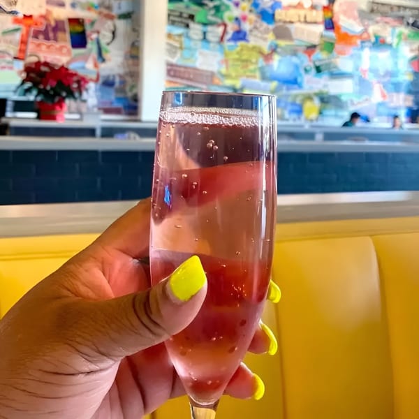 A hand with bright yellow nails holds a glass of pink sparkling drink in a vibrant restaurant.