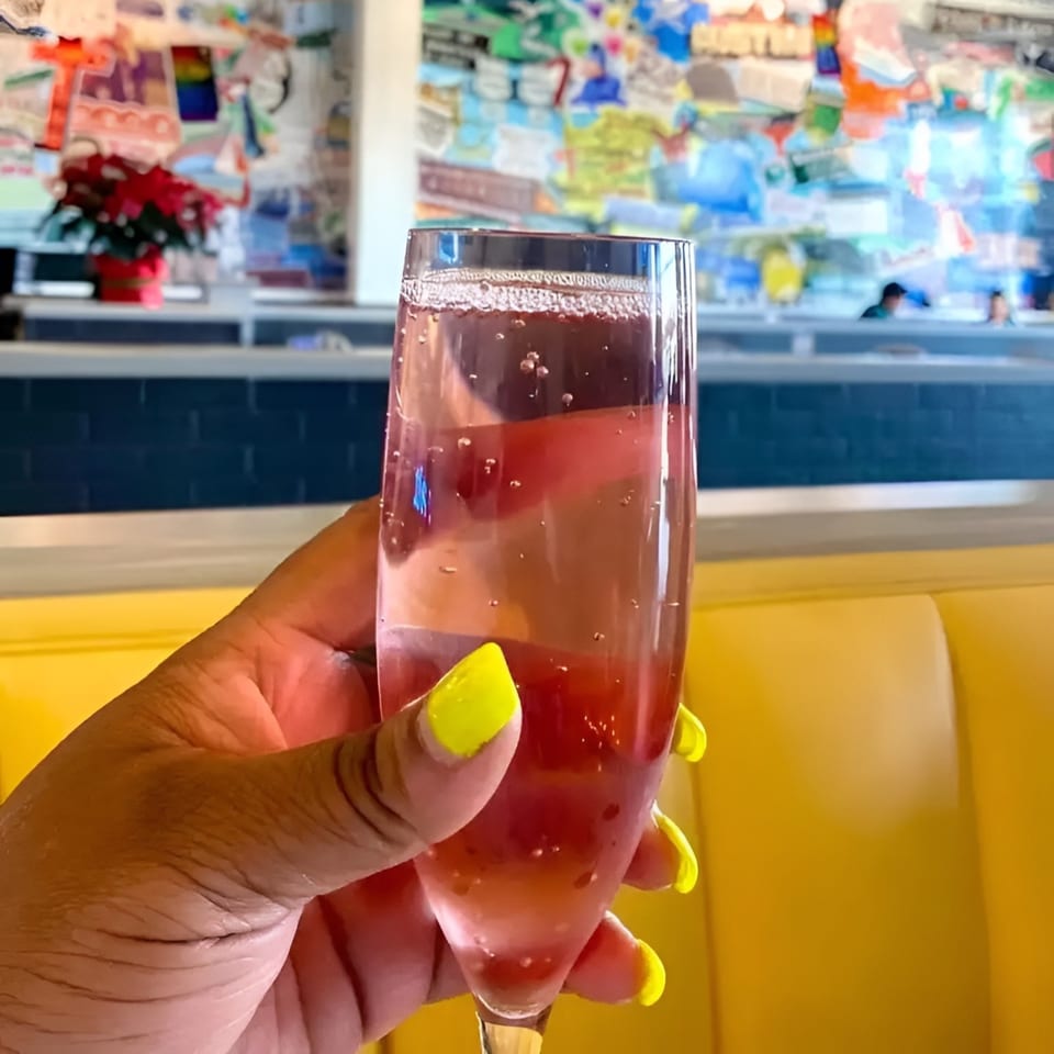 A hand with bright yellow nails holds a glass of pink sparkling drink in a vibrant restaurant.