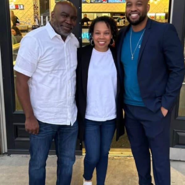 Three smiling people, two men and one woman, stand together in front of a restaurant.