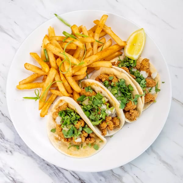 Three tacos with fried chicken, cilantro, and onions, served with french fries and a lemon wedge.