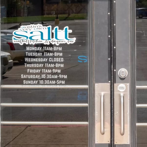 The front door of Saltt Seafood Kitchen & Daiquiri Bar, displaying its operating hours.