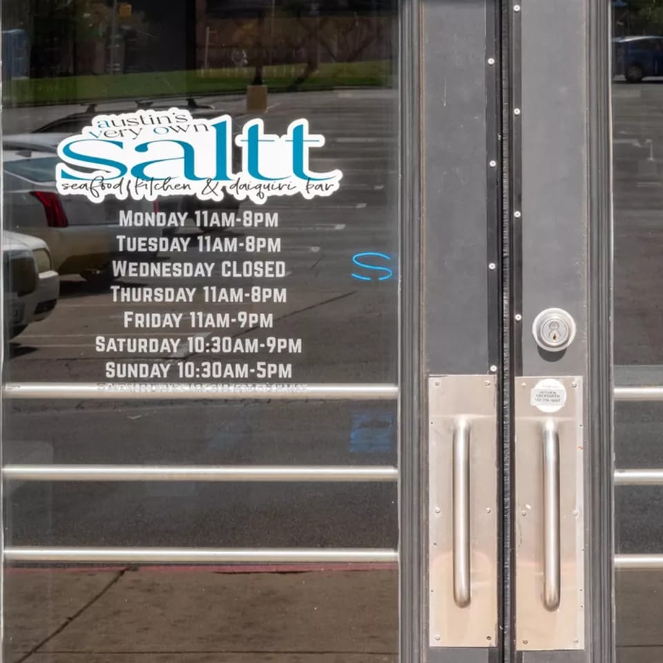 The front door of Saltt Seafood Kitchen & Daiquiri Bar, displaying its operating hours.
