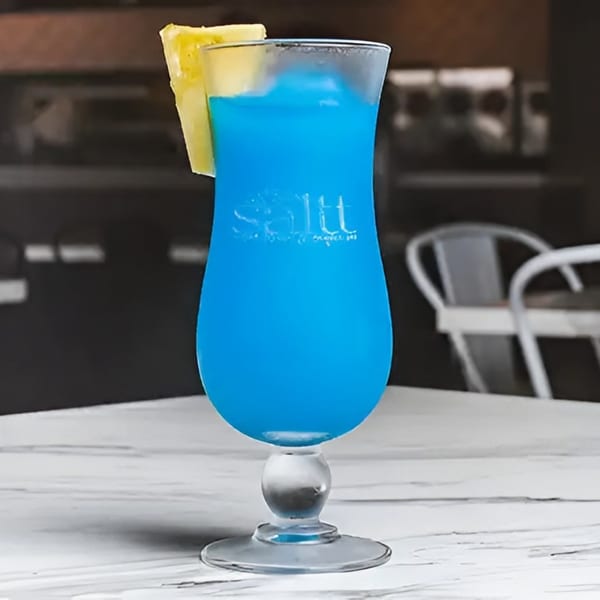 A vibrant blue cocktail in a hurricane glass, garnished with a pineapple slice, on a white marble table.