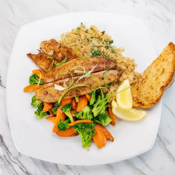 Seasoned fish fillets with rice, steamed broccoli and carrots, lemon wedges, and garlic bread.
