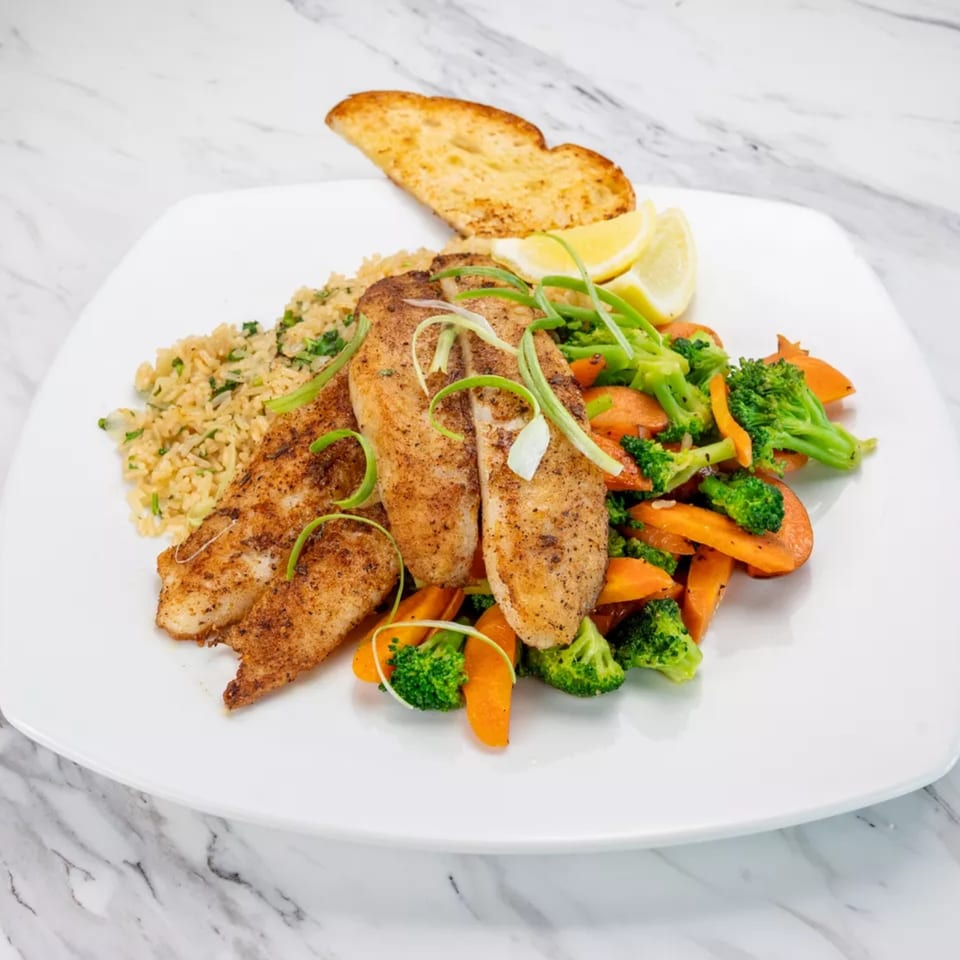Seasoned fish fillets with rice, mixed vegetables, lemon wedges, and garlic bread on a white plate.
