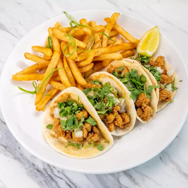 Three crispy chicken tacos with cilantro and onions, served with a side of french fries and a lemon wedge.