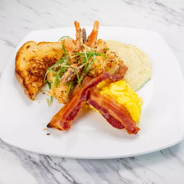 A plate of shrimp and grits with scrambled eggs, bacon, and toast on a white marble surface.