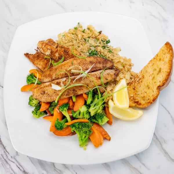 Seasoned fish fillets with rice, steamed broccoli and carrots, lemon wedges, and garlic bread.