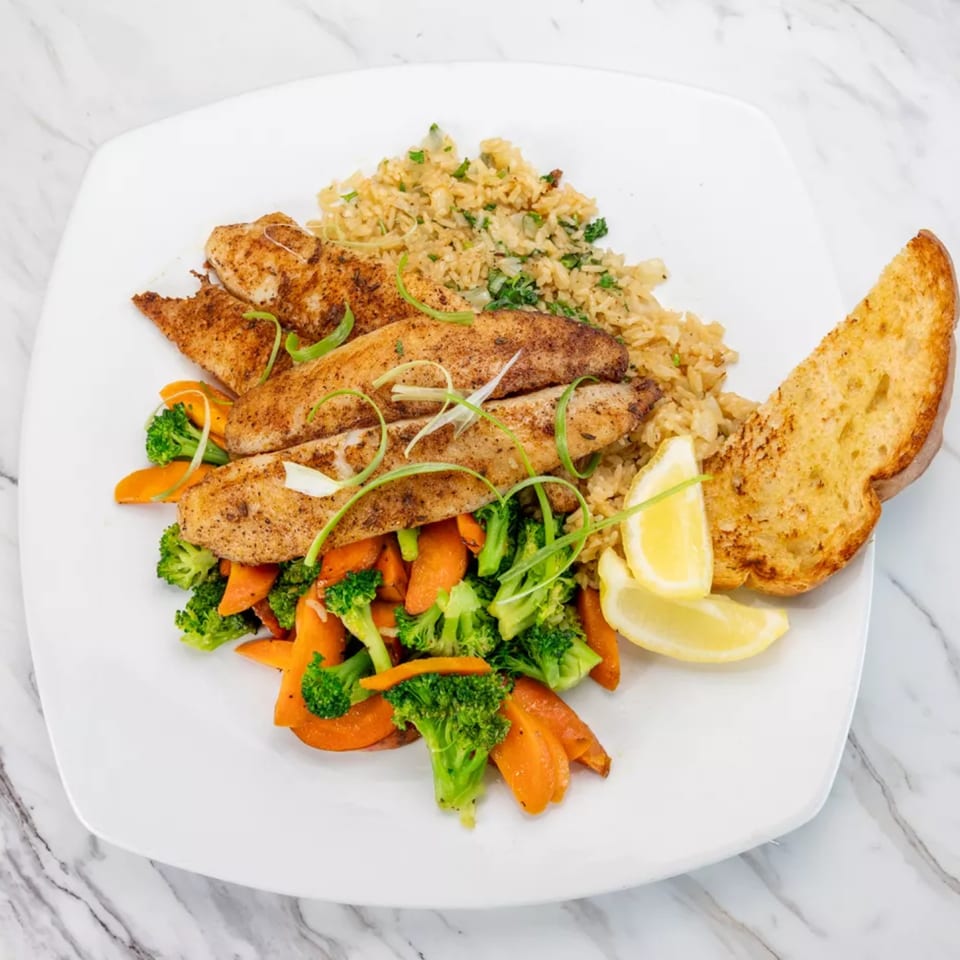 Seasoned fish fillets with rice, steamed broccoli and carrots, lemon wedges, and garlic bread.