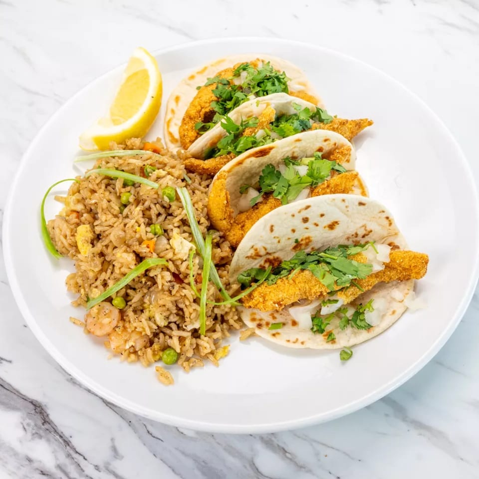 Three fish tacos with cilantro and onions, served with fried rice and a lemon wedge.