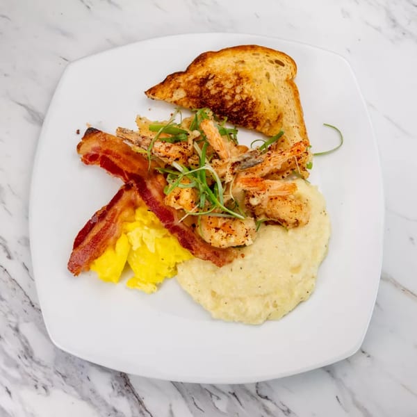 Shrimp and grits with scrambled eggs, bacon, and toasted bread on a white plate.