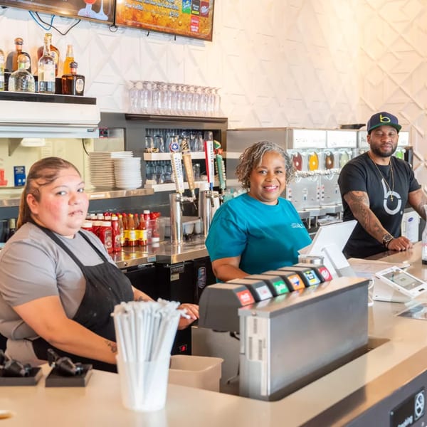 Three diverse restaurant staff members smiling behind a busy counter with drinks and equipment.