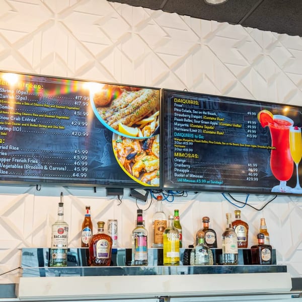 Restaurant menu boards display entrees, daiquiris, and mimosas above a bar shelf with liquor bottles.