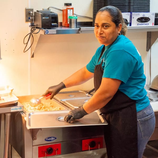 A woman in a blue shirt and black apron serves food from a hot bar in a kitchen.