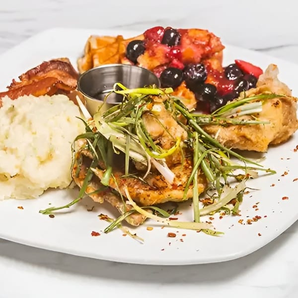 A plate with chicken, mashed potatoes, bacon, and waffles topped with berries and syrup.