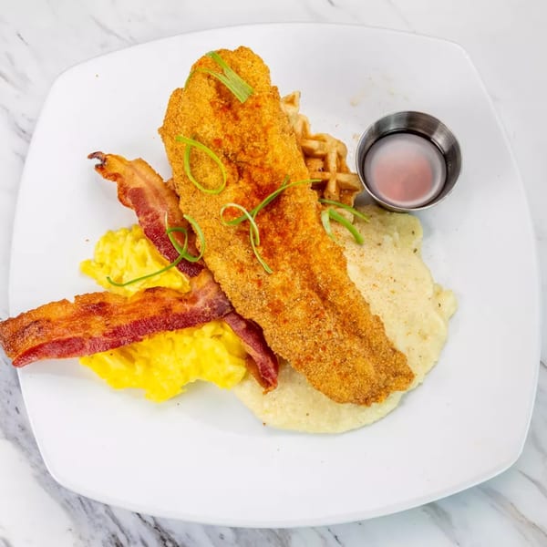 Fried fish with grits, scrambled eggs, bacon, a waffle, and syrup on a white plate.